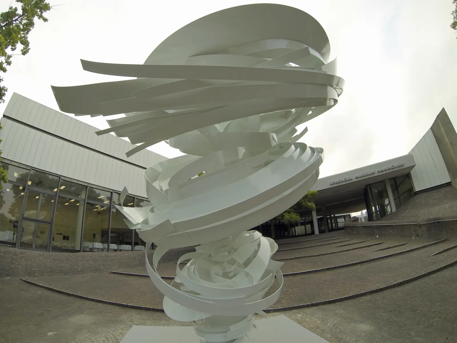 Weiße Stahlskulptur mit spiralförmigen Elementen vor einem Gebäude.