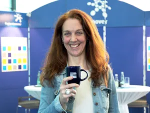 Eine Frau mit langen, roten Haaren steht l&auml;chelnd vor zwei Stehtischen und einer blauen Ausstellungswand. In der rechten Hand h&auml;lt sie eine Tasse mit dem Logo der Matinee im Foyer auf Brusth&ouml;he vor sich.