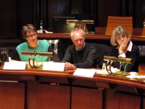 Drei Personen - zwei Frauen und ein Mann - sitzen im Hodlersaal in einer Sitzreihe hinter Pulten und blicken nach rechts. Auf den Pulten vor ihnen stehen Namensschilder.