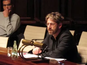 In der Mitte des Bildes sitzt eine Person mit Brille und Bart in einer Sitzreihe im Hodlersaal und spricht. Links oben im Hintergrund sitzt eine weitere Person und h&ouml;rt ihm zu.