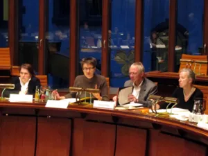 Vier Personen sitzen in einer nach innen gebogenen Reihe hinter einem langen Pult im Holdersaal. Jeschke hebt den Zeigefinger und spricht, T. Walter schaut in einen Aktenordner auf dem Tisch vor ihm. Auf den Tischen stehen Namensschilder.
