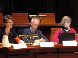 Drei Personen sitzen im Holdersaal hinter Pulten, vor ihnen steht ein Laptop. Zwei schauen nach vorne, die mittlere Person sieht nach unten.