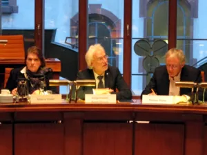 Drei Personen sitzen hinter Pulten im Hodlersaal. Die linke Person schaut nach vorne, die Mittlere spricht und schau dabei nach rechts. Die Rechte Person blickt auf ein Papier auf dem Pult vor sich und h&auml;lt einen Stift in der Hand.