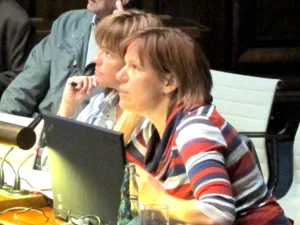 Eine Frau sitzt hinter einem aufgeklappten Notebook und schaut nach vorne. Hinter ihr sind drei weitere sitzende Personen erkennbar.