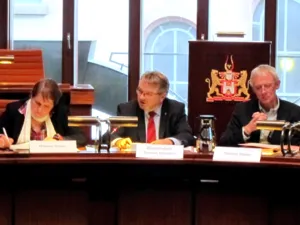 Drei Personen sitzen im Hodlersaal nebeneinander hinter Pulten. Die mittlere schaut zur Seite und spricht.