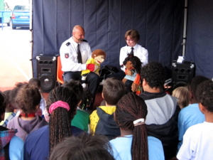 Etwa 20 Kinder sitzen in einem Zelt. Vor ihnen sitzen eine Polizist und eine Polizistin mit Handpuppen.