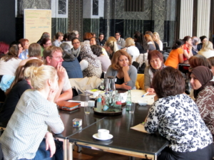 Acht Personen sitzen um einen Tisch herum und sprechen miteinander. Im Hintergrund sind die drei weiteren Gruppen erkennbar.
