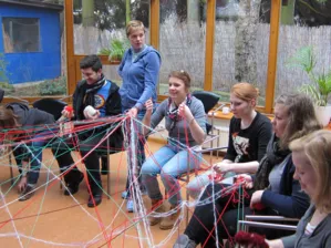 Eine Gruppe von Jugendlichen sitzt im Kreis, wobei jeder von Ihnen untereinander gespannte F&auml;den in der Hand h&auml;lt