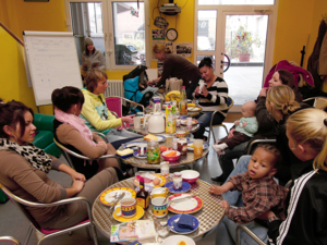 Mehrere junge Frauen sitzen mit ihren kleinen Kindern an Tischen, auf denen Frühstücksutensilien stehen