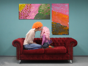 Frau und Kind mit Perücken auf einem Sofa. An der Wand dahinter hängen abstrakte Bilder, deren Farbgebung mit den Perücken harmoniert.