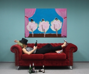 Eine Frau liegt auf einem Sofa mit einer Sektflasche in der Hand.Über ihr hängt ein Bild an der Wand, das Frauen beim Can-Can-Tanzen zeigt.