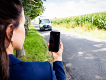 Eine Frau steht im Freien an einer Straße und schaut auf ihr Handy. Ein Transporter nähert sich.