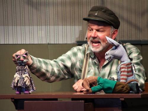 Mann mit einer Katzenpuppe, einer Eselpuppe und einer Hundepuppe.