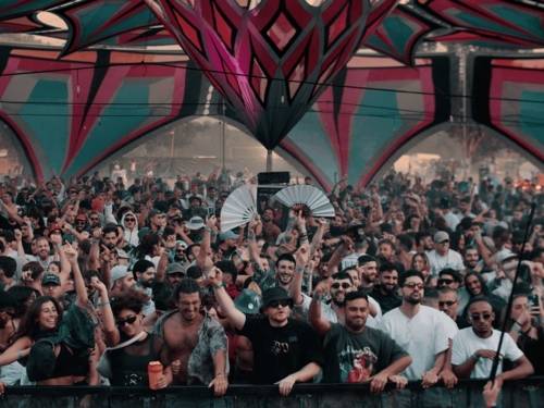 Eine große Menschenmenge feiert vor einer Bühne unter einem farbenfrohen, geometrisch gestalteten Zeltdach. Die dicht gedrängten Festivalbesucherinnen und Festivalbesucher tanzen, heben die Arme in die Luft und schaffen eine lebendige, energiegeladene Atmosphäre.