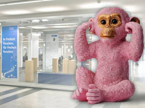 Große rosafarbene Puppe eines Affen, der sich die Ohren zuhält vor einem Plakat mit der Aufschrift "In Freiheit forschen, für Freiheit forschen".