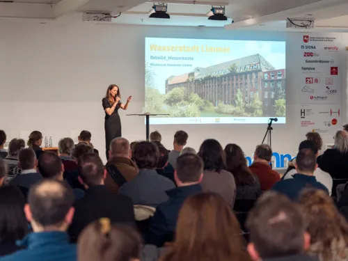 Ein Publikum von hinten sieht auf eine Plattform, auf der eine Frau an einem Stehtisch mit Mikrofonen steht, rechts daneben eine auf die Wand projezierte Seite mit "Wasserstadt Limmer. Rebuilt_Wasserkante".