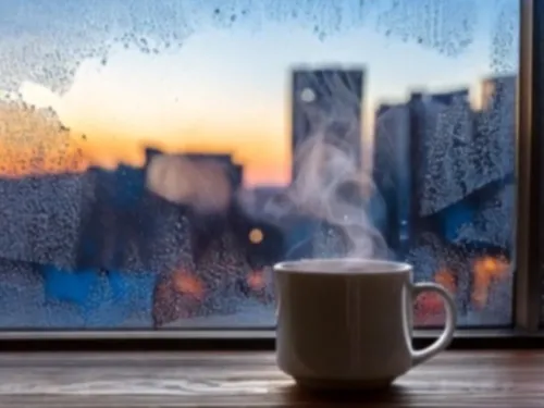 Ein Fenster mit Eis, dahinter eine Stadt mit Gebäuden, vorn ein Becher mit dampfendem Kaffee.
