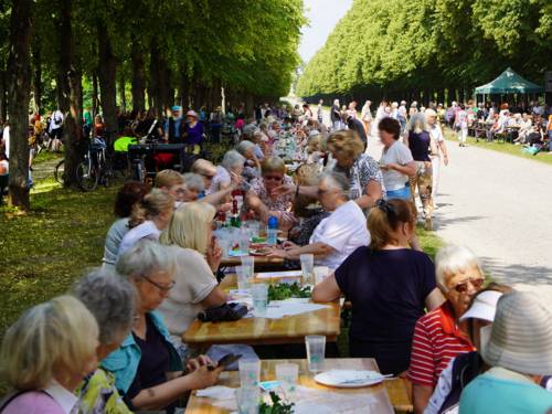 Durch eine lange Baumallee läuft ein Weg, darauf steht eine lange Tafel mit vielen Menschen daran, überwiegend ältere Menschen. Sie unterhalten sich und essen.