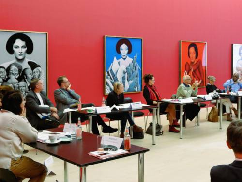 Elf Personen (5 Frauen und 3 Männer) sitzen in einem rot gestrichenen Raum an Tischen und schauen zur rechten Bildseite. Dort scheint ein Mann einen Redebeitrag zu halten. Auf den Tischen sind Arbeitsmaterialien, Namensschilder und Getränke zu sehen.