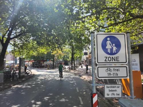 Eine Straße mit Radfahrer und Bäumen, rechts ein Schild Fußgängerzone