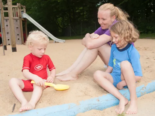 Eine erwachsene Frau sitzt im Sand und schaut gemeinsam mit einem Mädchen zu, wie ein kleiner Junge mit einer Schaufel buddelt.