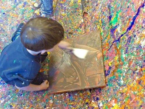Ein Kind bemalt eine Leinwand und hockt dabei auf bunt besprenkeltem Malervlies