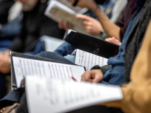 Menschen sitzen in Stuhlreihen nebeneinander und haben Mappen mit Noten auf dem Schoß.
