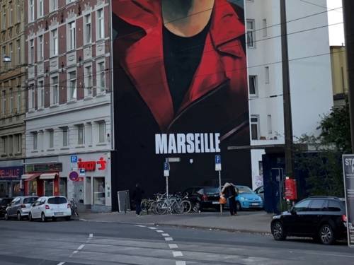 Sichtbar ist ein Häuserblock dessen Seite mit einem Portraitbild eines Mannes bemalt ist. Das Portrait stellt den Charakter "Marseille" aus der Serie Haus des Geldes dar.