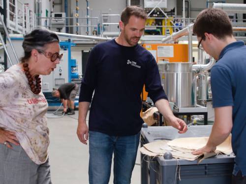 Eine Frau und zwei Männer in Fabrik über einen Tisch gebeugt.