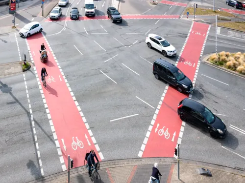 Eine Kreuzung von oben mit roten Radwegen, darauf Radfahrende, Stra&szlig;e und Autos.