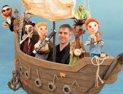 Ein Mann und fünf Puppen mit Instrumenten sitzen in einem hölzernen Segelboot.