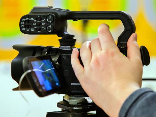 Eine Filmkamera filmt auf einem Stativ, die Hand einer Person ist an der Kamera.