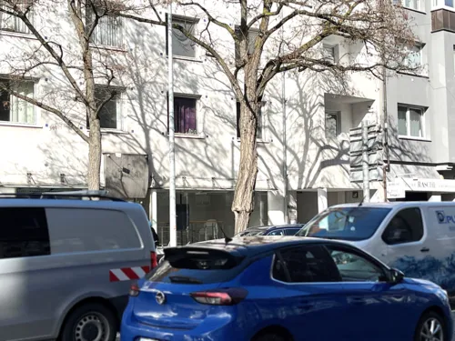Drei Autos dicht aneinander im Verkehr auf einer Straße vor einem weißen Wohnhaus, dazwischen ein kahler Baum.