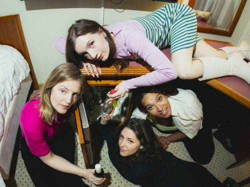 Vier Frauen belagern den Mini-Kühlschrank eines Hotelzimmers und schauen in die Kamera.