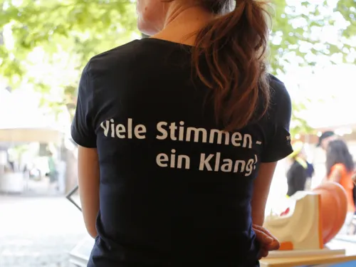 Eine von hinten fotografierte Person. Auf dem T-Shirt steht "Viele Stimmen, ein Klang!"