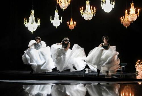 Drei Frauen tanzen wild in weißen Ballkleidern auf einer schwarz gehaltenen Bühne, bei der Kronleuchter von der Decke hängen.