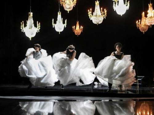 Drei Frauen tanzen wild in weißen Ballkleidern auf einer schwarz gehaltenen Bühne, bei der Kronleuchter von der Decke hängen.