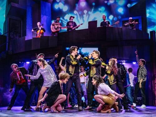 Zu sehen ist eine Performance auf der Bühne mit großem Ensemble zum Musical „Abenteuerland“. Auf einer oberen Ebene erkennt man Teile eines Orchesters.