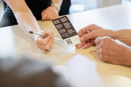 Ein Tisch mit den Händen von zwei Personen. Eine Person hält einen Flyer, die andere Zeigt mit einem Stift auf einen QR-Code darauf.