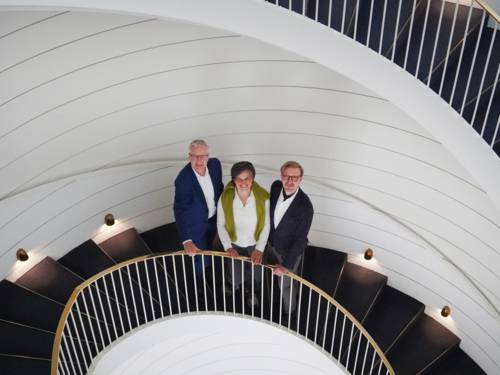 Blick von oben auf eine kreisrunde dunkle Treppe vor weißer Wand und mit weiß-goldenem Geländer, die sich wie ein Schneckenhaus windet. Auf der Treppe stehen eine Frau und zwei Männer.