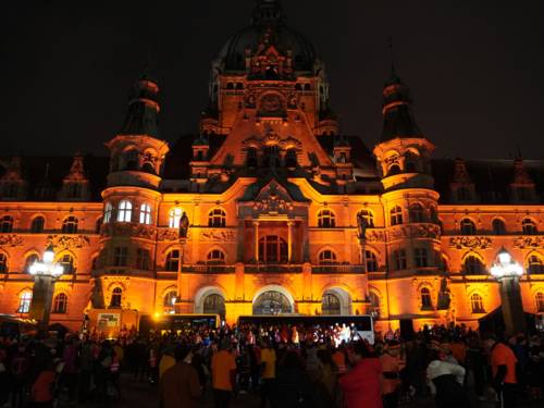 Ein altes schlossähnliches Gebäude im Dunkeln im orangem Licht angestrahlt. Davor stehen einige Menschen.