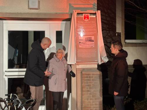 Zwei Männer und eine ältere Dame stehen vor einem Haus, an der Wand eine Tafel mit einem kleinen geöffneten Vorhang.