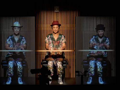 Eine Frau sitzt auf einer Bühne und spielt ein imaginäres Klavier, links und rechts von ihr sind lebensgroße Videoproduktion von ihr selbst.