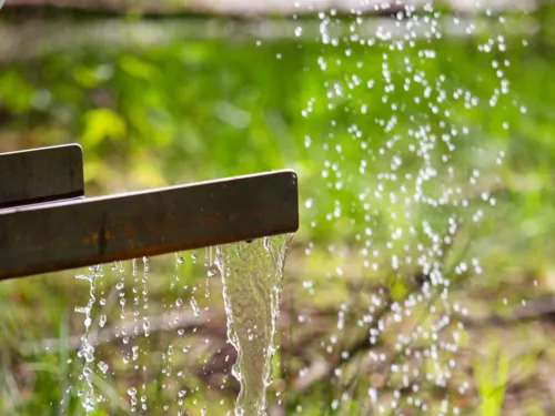 Wasser fließt Über eine Rinne und fällt auf bewachsenen Boden.