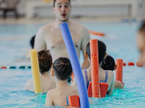 Schwimmlehrer und Kinder mit Poolnudeln und anderen Auftriebshilfen