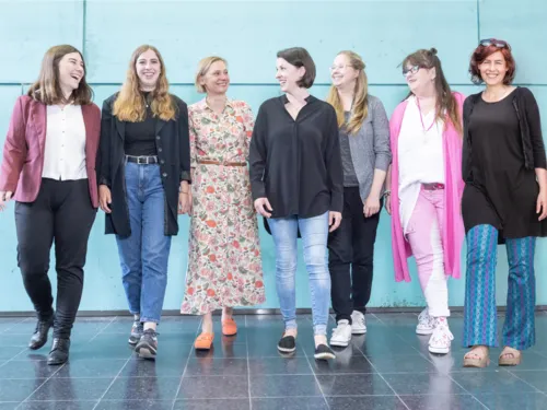 Sieben Frauen lachen und gehen auf die Kamera zu, im Hintergrund ist eine türkisfarbene Fassade.