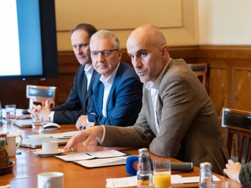 Drei Männer sitzen nebeneinander an einem Tisch, leicht seitlich dargestellt. Der vordere spricht.