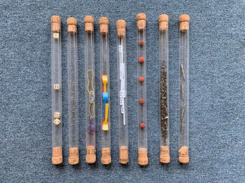 Acht durchsichtige Röhrchen liegen nebeneinander auf einem Teppich. Jedes Röhrchen ist an beiden Enden mit jeweils einem Korken verschlossen. In den Würfeln sind (von links nach rechts): 1.: drei Würfel mit Zahlen, 2.: drei Büroklammern, 3. fünf Gummiringe, 4.: fünf Speiseeislöffel, 5.: acht Kaffeeumrührstäbchen, 6.: fünf rote Kügelchen, 7.: sehr viele Sonnenblumenkerne, die dieses Röhrchen zu ca. zwei Dritteln füllen und 8.: vier Metallzapfen.