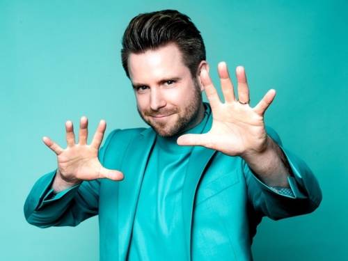 Zu sehen ist ein Mann mit dunklem, nach hinten gegelltem Haar. Er trägt einen Türkisfarbenen Anzug und T-Shirt und steht vor einem einfarbigen Hintergrund derselben Farbe. Seine sind in typischer Magier Pose nach vorne gerichtet.