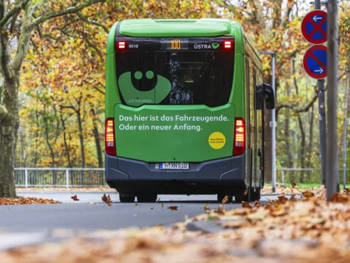 Ein eCitaro fährt mit dem Heck in Richtung Kamera auf einer von Bäumen umrandeten Straße. Auf dem Heck ist ein großes ÜMO und der Spruch "Das hier ist das Fahrzeugende. Oder ein neuer Anfang." Es liegt Laub am Straßenrand. Im Hintergrund ist der weitere Straßenverlauf.